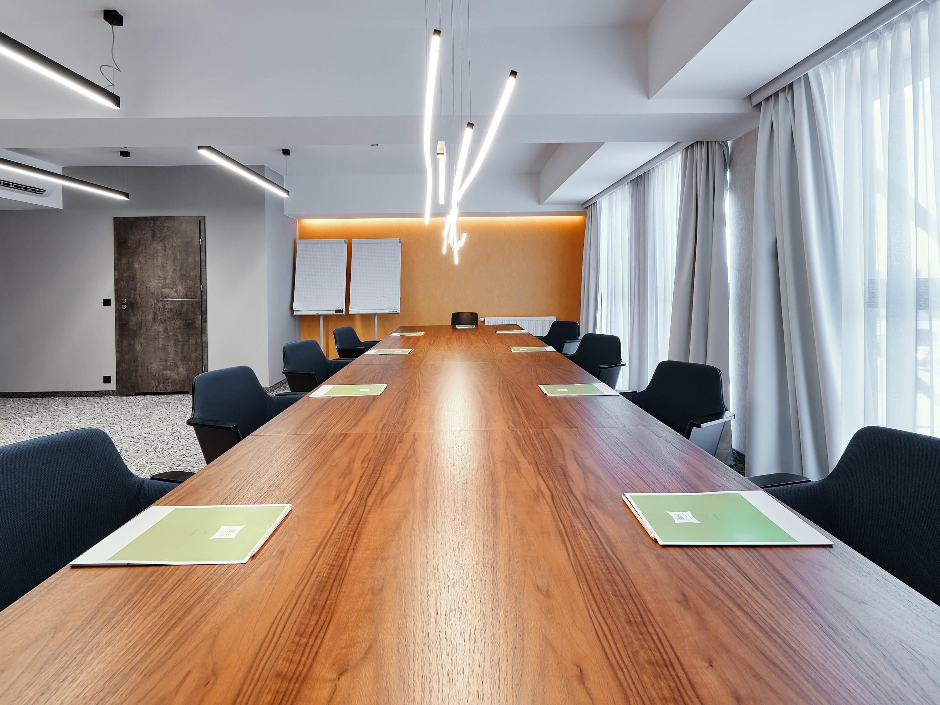 A conference room with a long wooden table, black chairs, white curtains, and a whiteboard.
