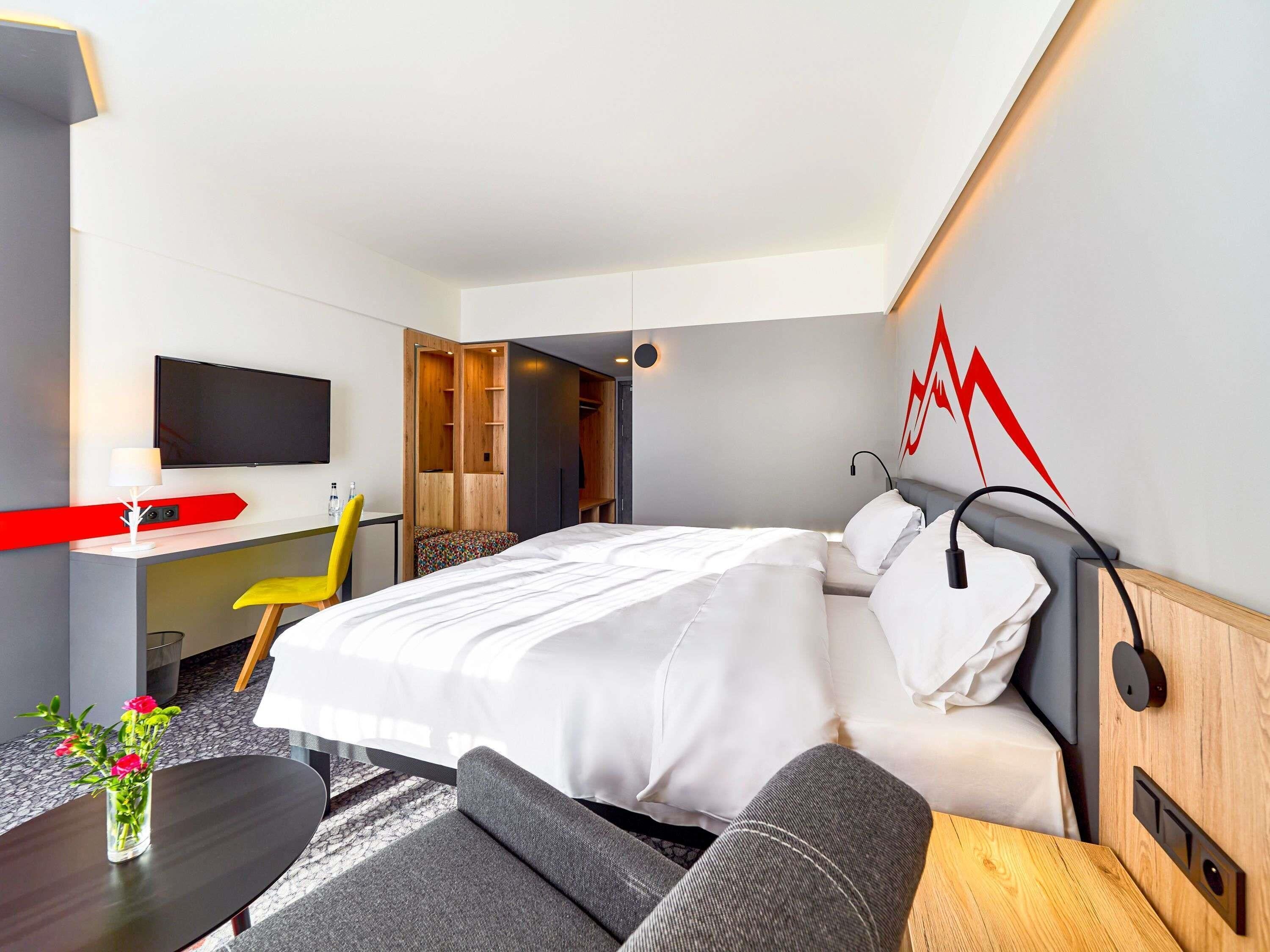 Una habitación de hotel moderna con cama, escritorio, silla, televisión y un elemento decorativo rojo.