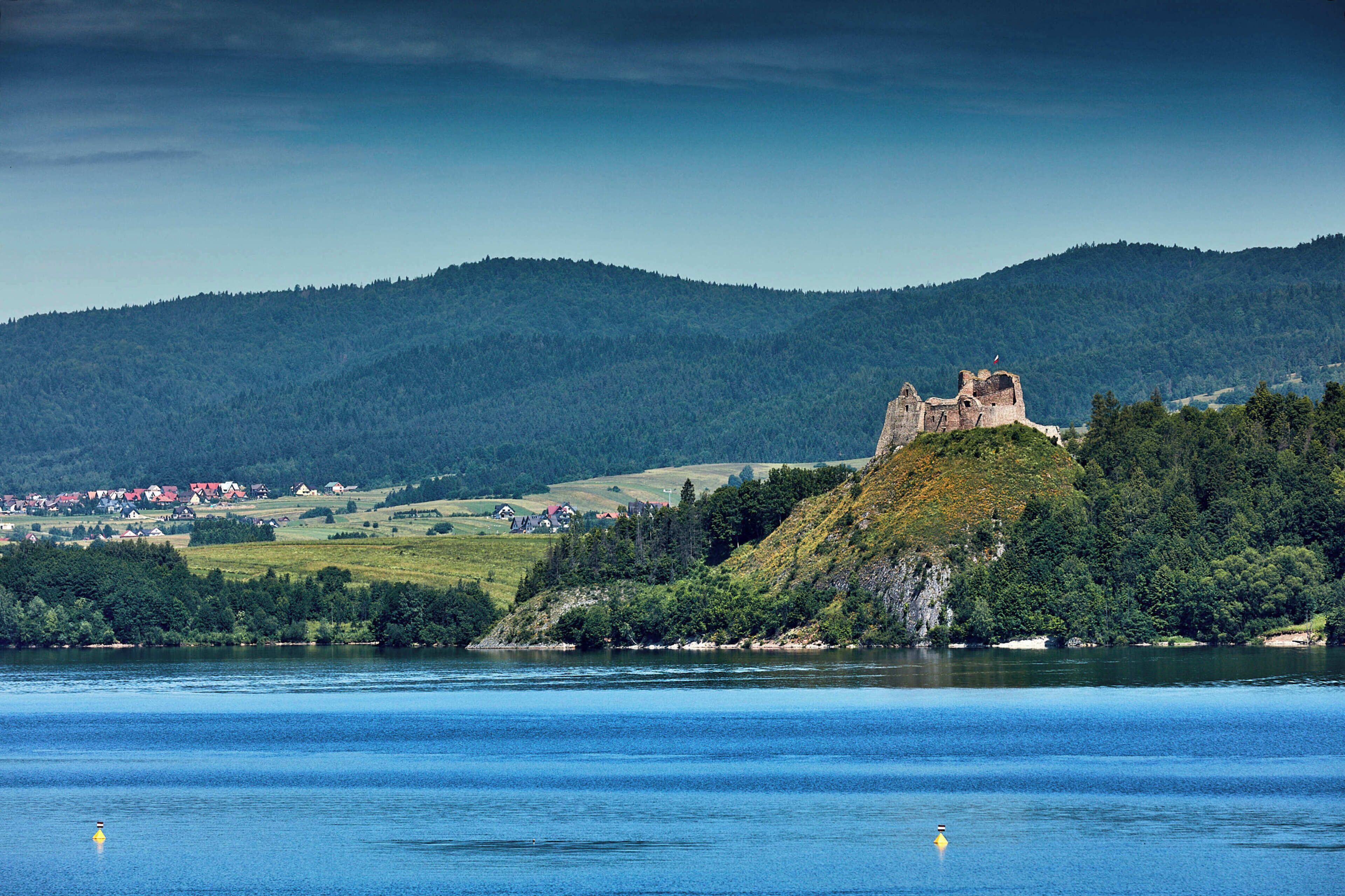 A castle on a hill overlooking a body of water with a forested landscape in the background.