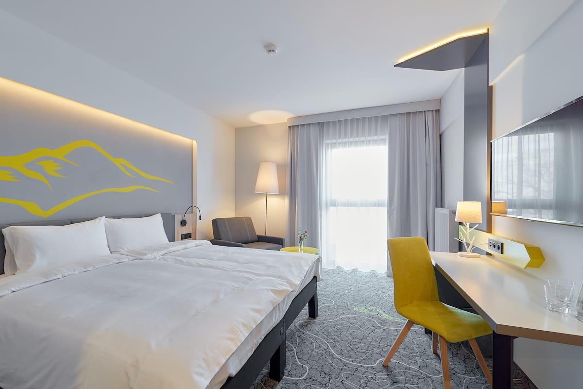 A modern hotel room with a large bed, a desk, a chair, and a window with curtains.