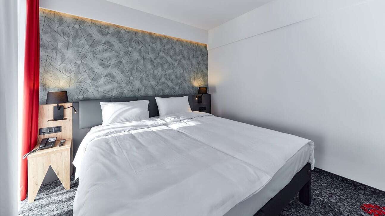 Et moderne hotelværelse med en stor seng, en sengegavl med et bladmotiv, sengeborde med lamper og en rød gardin.