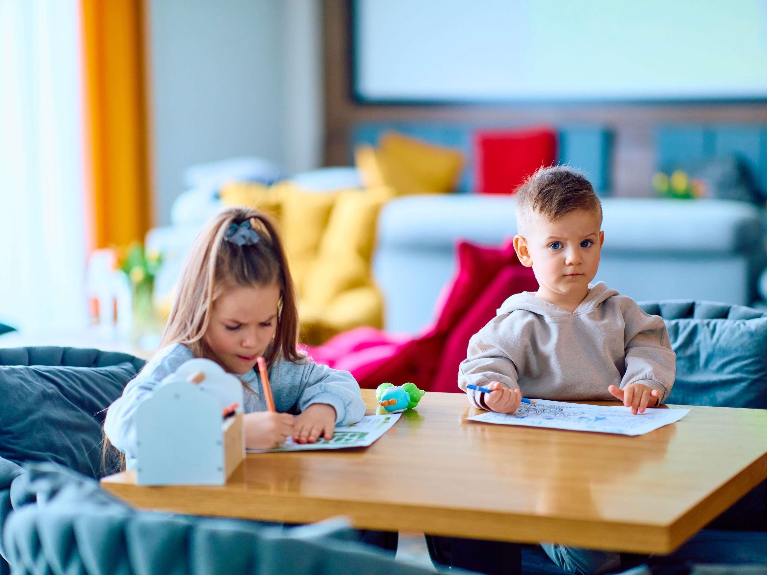 Two children sitting at a wooden table with papers, in a room with a sofa and cushions.