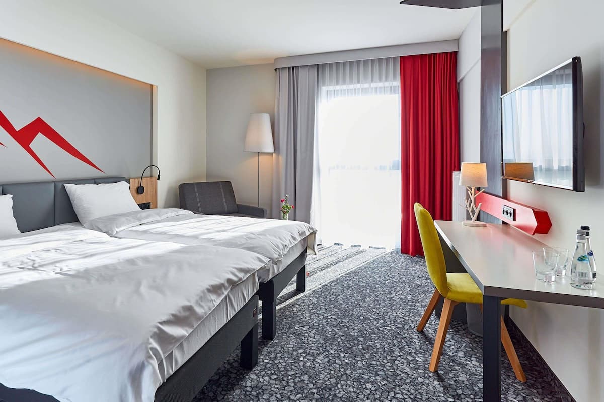 A hotel room with a bed, a desk, a chair, a lamp, a TV, and a window with curtains.