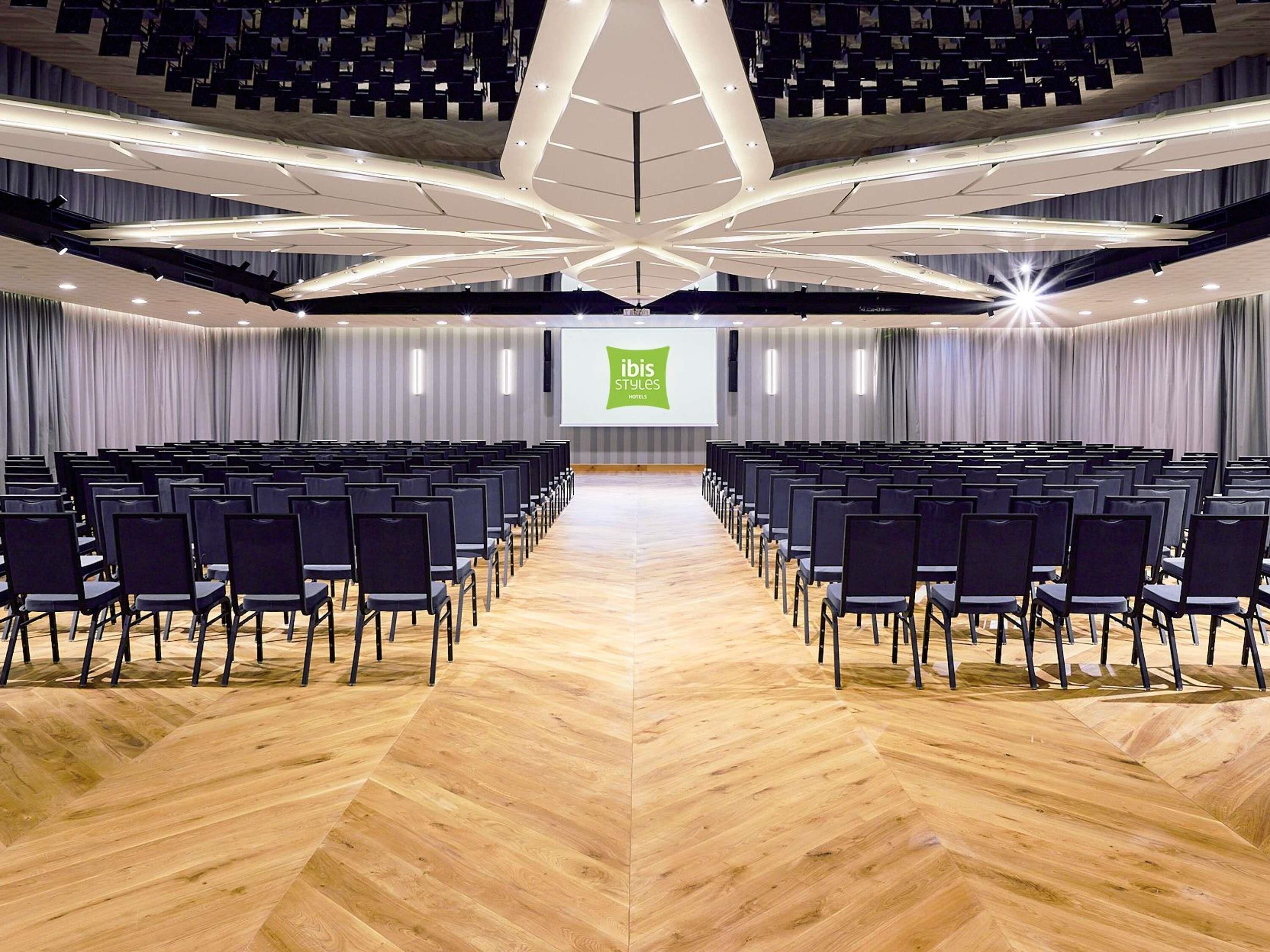 A conference room with rows of chairs, a stage with a presentation screen, and a modern ceiling design.