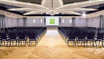 A conference room with rows of chairs, a stage with a presentation screen, and a modern ceiling design.