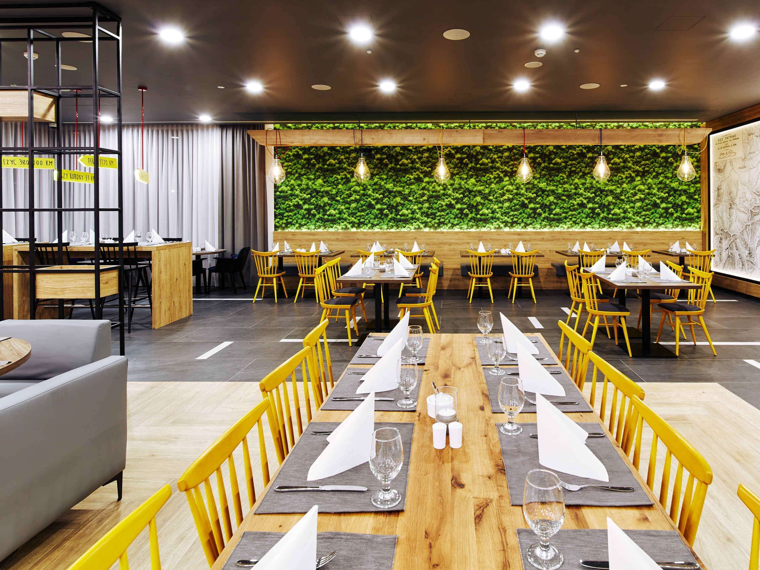 A restaurant with long wooden tables, yellow chairs, and a green wall with plants.