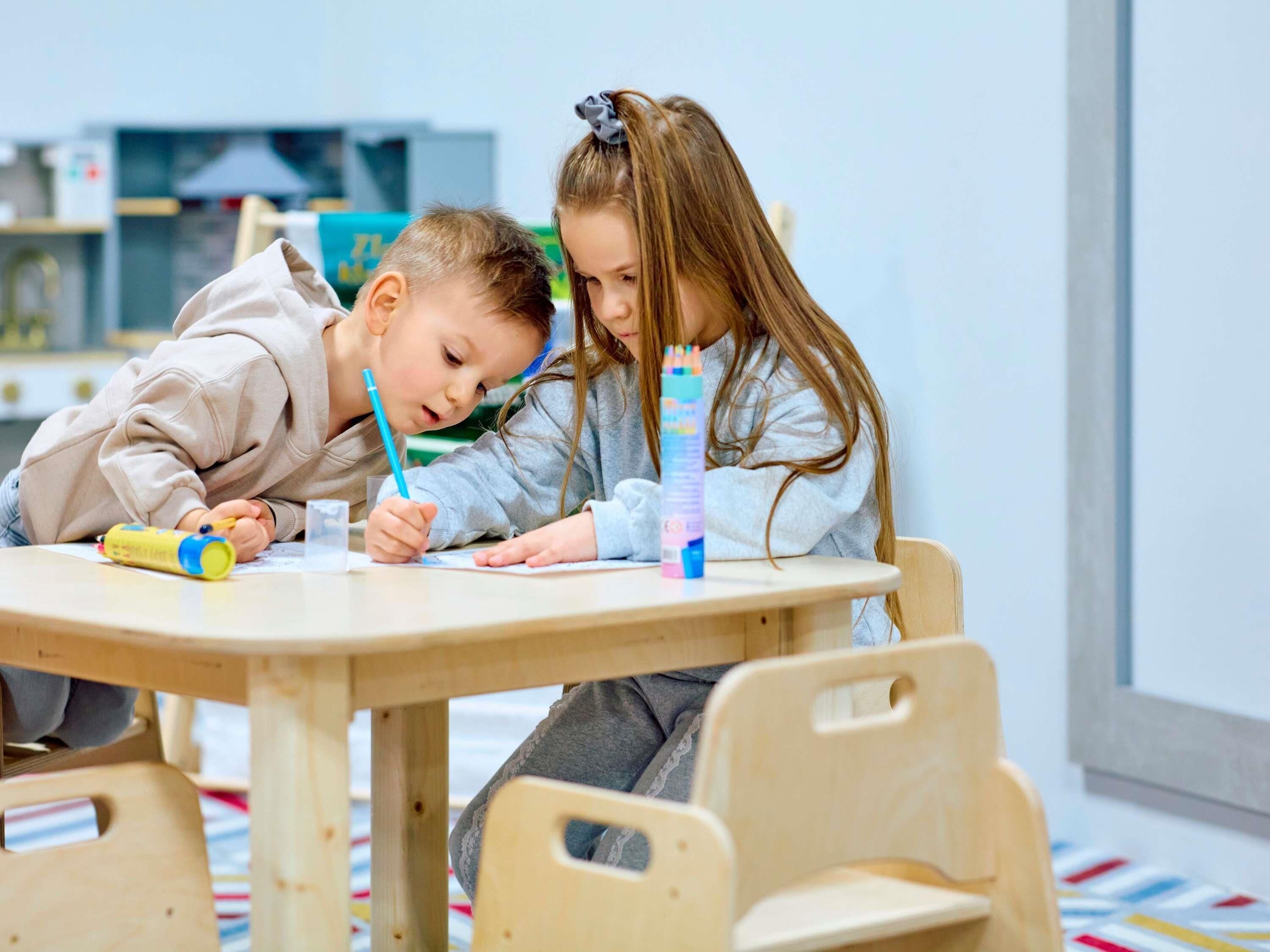 Dos niños en una mesa de madera con sillas, escribiendo y dibujando, en una habitación con una ventana y estanterías al fondo.