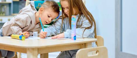 Dos niños en una mesa de madera con sillas, escribiendo y dibujando, en una habitación con una ventana y estanterías al fondo.