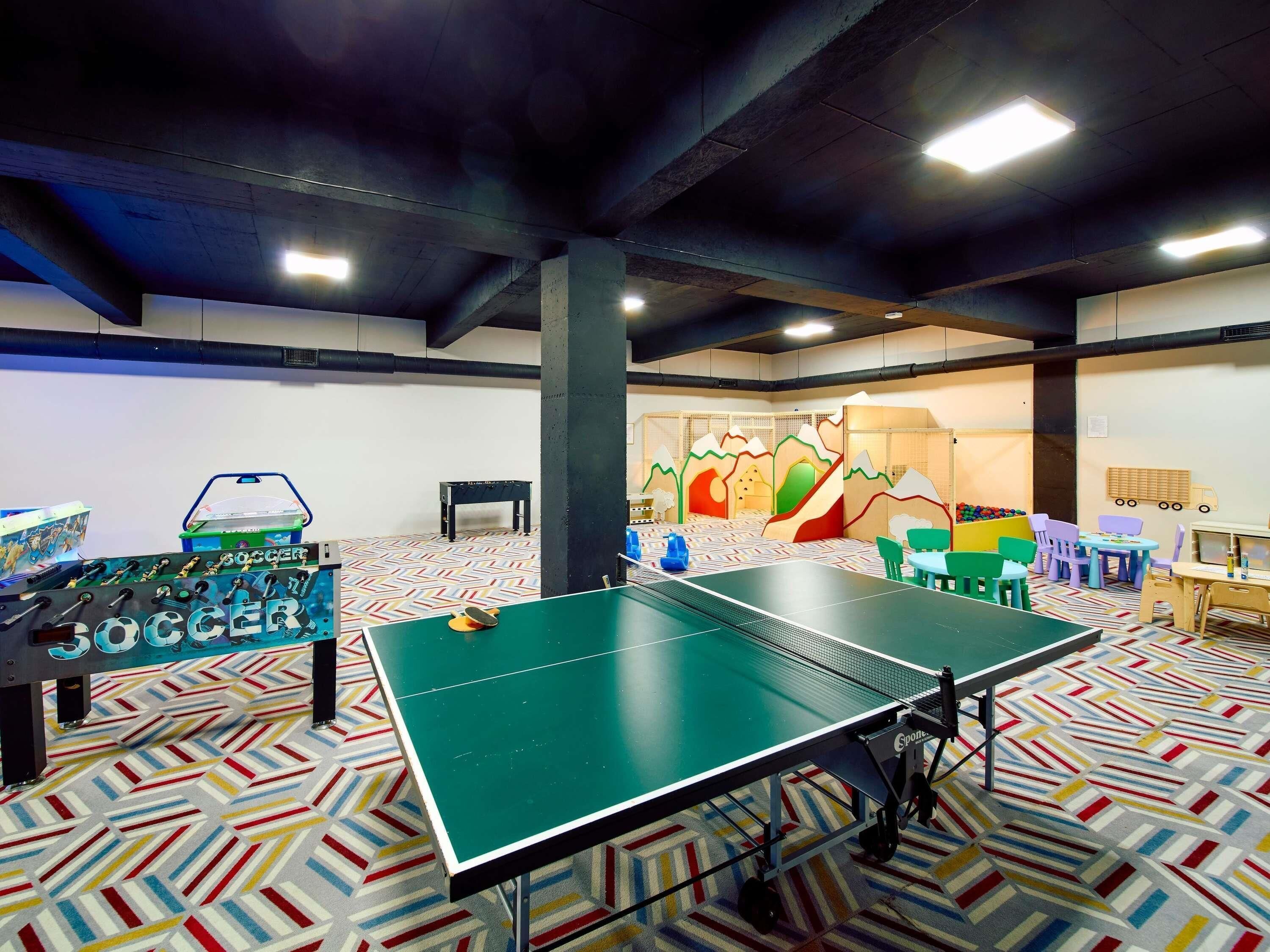 Una mesa de ping pong en una habitación con una alfombra geométrica de colores, una mesa de fútbol y una zona de juegos con un mural temático de montaña.