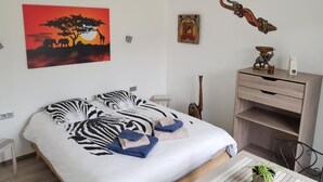 A bedroom with a bed, a desk, a chair, a potted plant, and wall art including a zebra print bedspread, a sunset painting, and a wooden sculpture. - Chambres d'hôtes de l'Orval (Thiergeville)