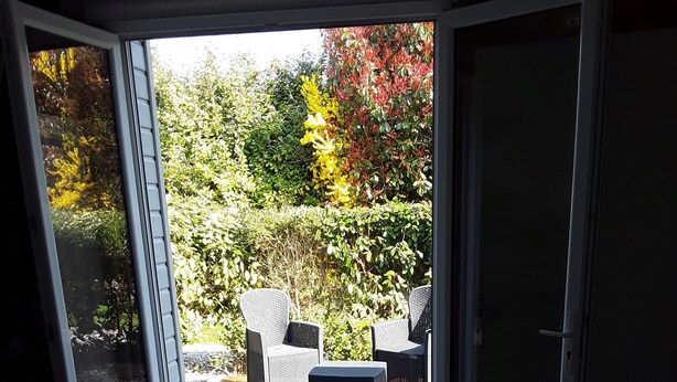 A room with a sliding glass door leading to an outdoor patio area with wicker furniture and a view of a garden. - Chambres d'hôtes de l'Orval (Thiergeville)
