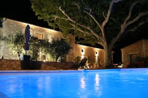 A nighttime view of a swimming pool area with a stone building, trees, and outdoor seating. - Le Mas de Nonin - Chambres d'hotes (Noves)