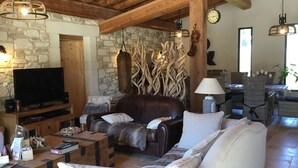 Un salon chaleureux avec un mur en pierre, des poutres apparentes, un canapé en cuir, un téléviseur et un coin repas avec des meubles en osier.