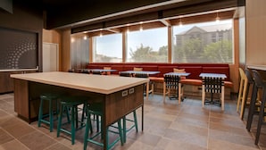 A modern dining area with a long table, multiple booths, and a large window providing a view of buildings and trees. - Tru by Hilton Grand Prairie (Grand Prairie)