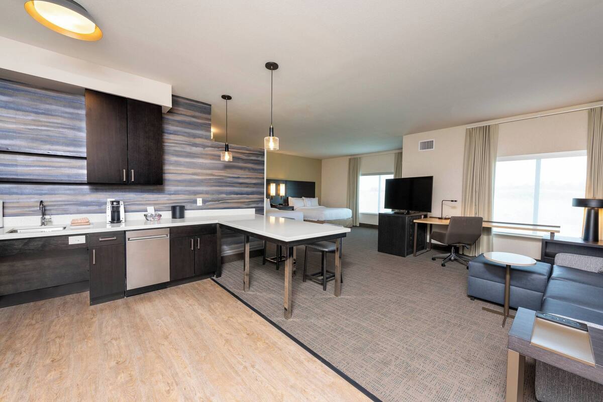 A modern hotel room with a kitchen area, a dining table, a sofa, and a TV.