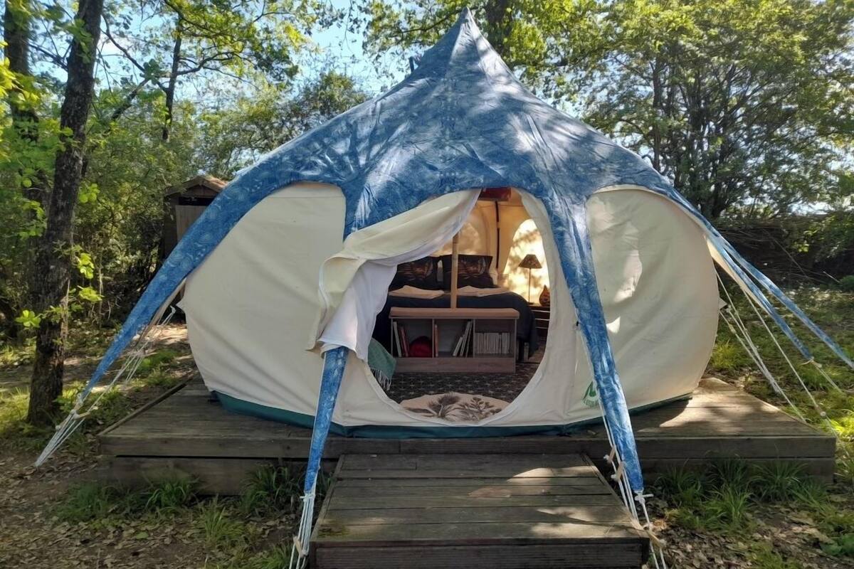 A tent with a wooden deck entrance, a bed inside, and a view of the surrounding forest.