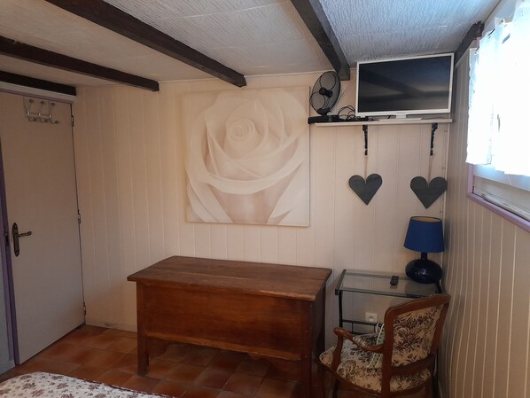 A room with a wooden table, a chair, a television on a shelf, and a large white rose painting on the wall. - Le Clos des Epinettes (Angé)