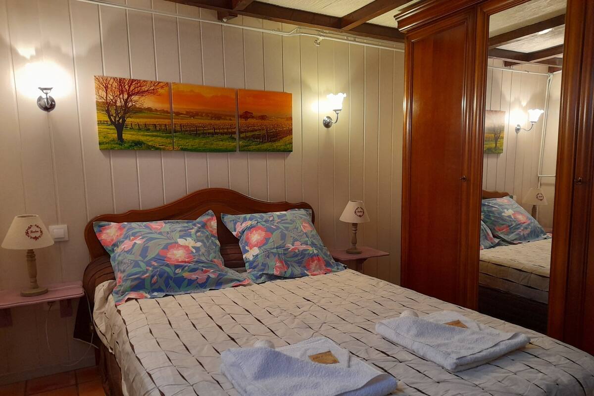 A neatly made bed with floral pillows and a quilt, a bedside lamp, a painting of a sunset on the wall, and a mirror reflecting the room.
