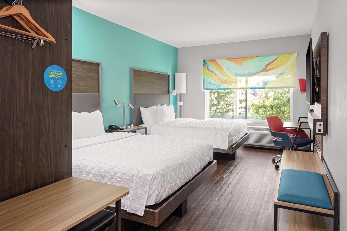 A hotel room with two beds, a desk, a chair, a TV, and a window with a view of greenery.