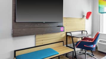 Een moderne kamer met een grote flatscreen televisie bevestigd aan een houten paneel, een bureau met een stoel, een bankje en een raam met een kleurrijk gordijn.