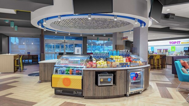 Un quiosco de aperitivos con diversas opciones y un refrigerador, ubicado en un espacio interior moderno con zona de estar y una tienda visible llamada 'TOPIT'.