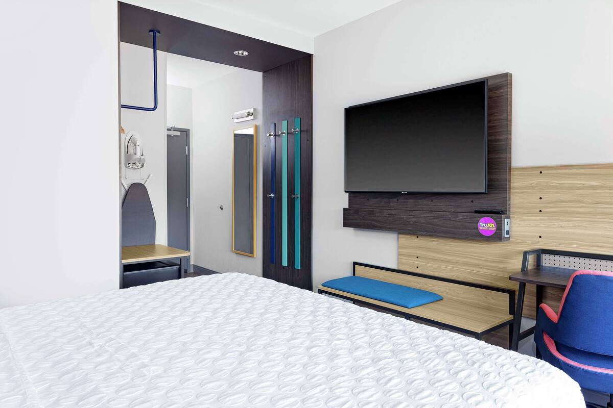 A modern hotel room with a large bed, a desk with a chair, a flat-screen TV mounted on the wall, and a mirror.