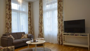 Een woonkamer met een bank, een salontafel, een televisie op een meubel en grote ramen met bloemen gordijnen.