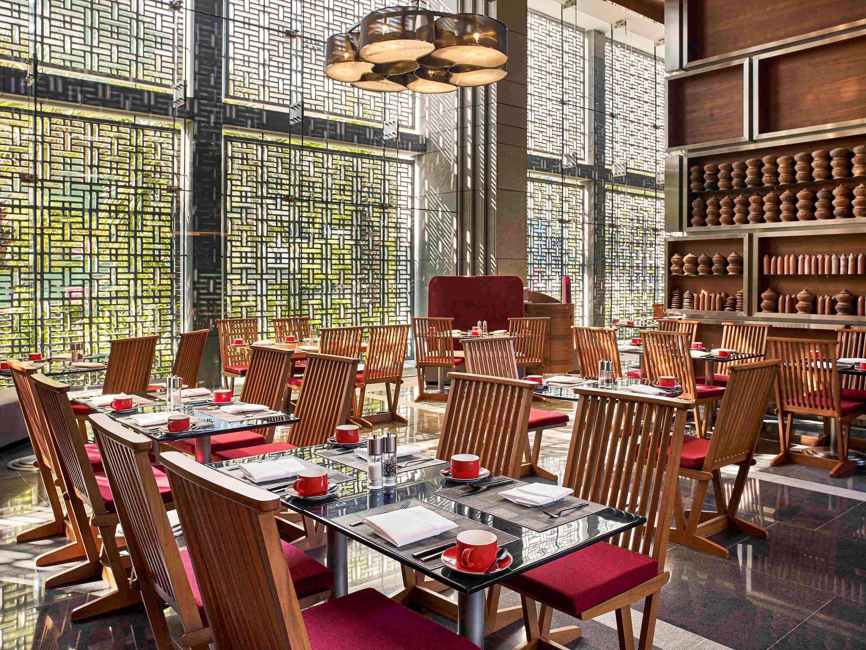 Un restaurante moderno con sillas y mesas de madera, ventanas grandes con un diseño geométrico y una estantería con cerámica.
