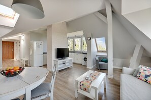 A spacious living area with a dining table, a sofa, and a TV. There is a kitchen with a washing machine and a window with a view of trees. - Grand Apartments - Love Loft (Sopot)