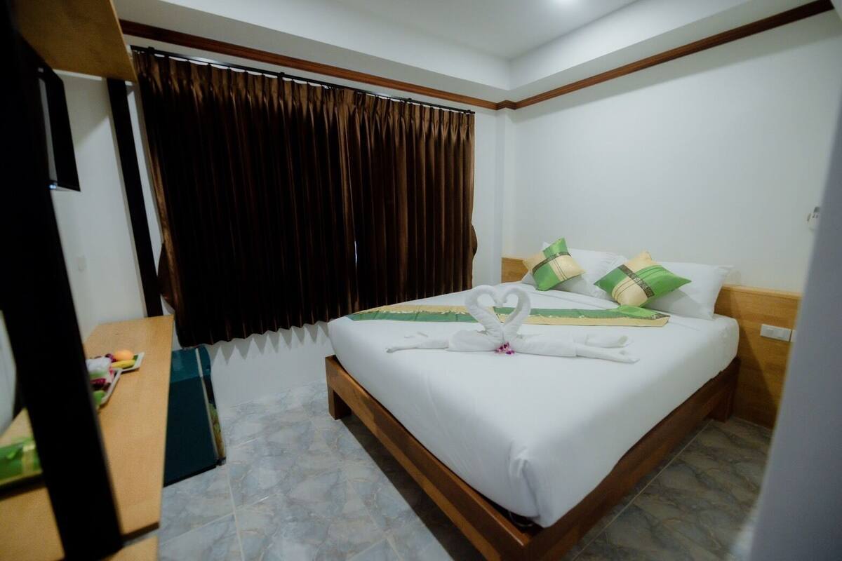 A hotel room with a neatly made bed, a wooden headboard, a window with dark curtains, and a small table with a fruit plate.