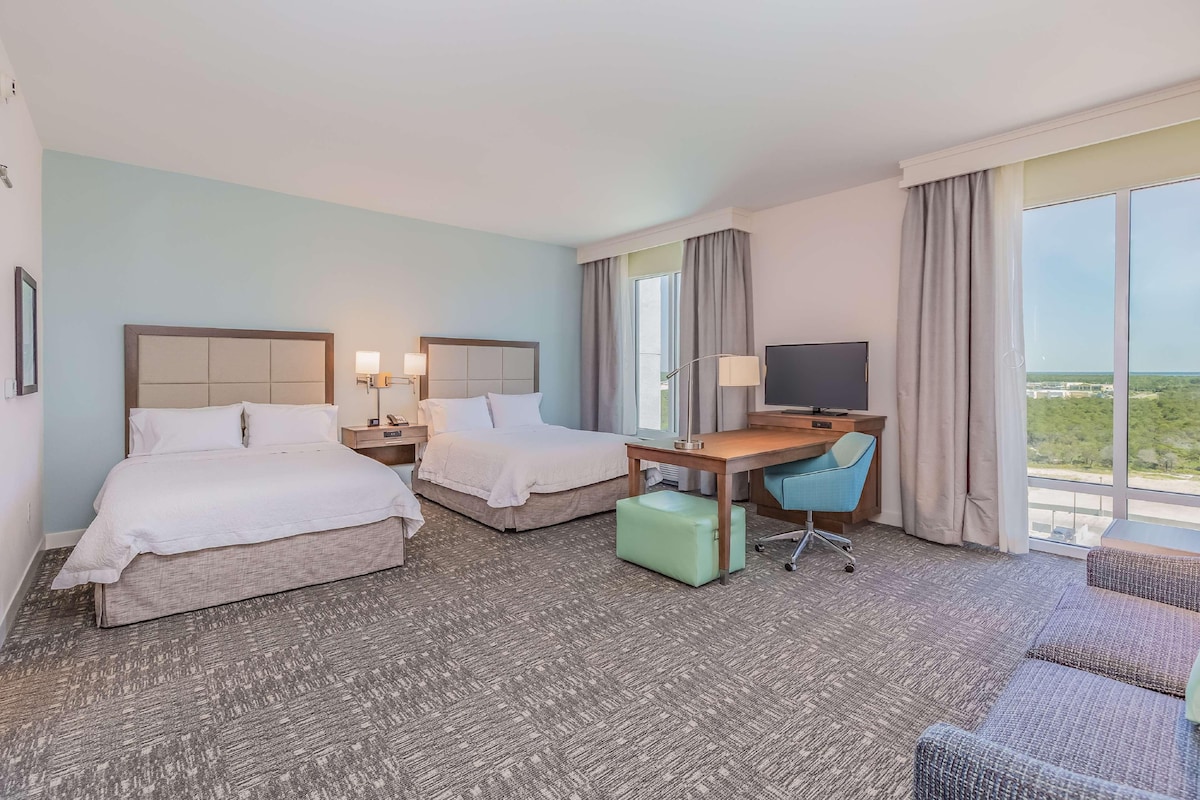 Habitación de hotel con dos camas, un escritorio, un televisor y un ventanal con vistas.