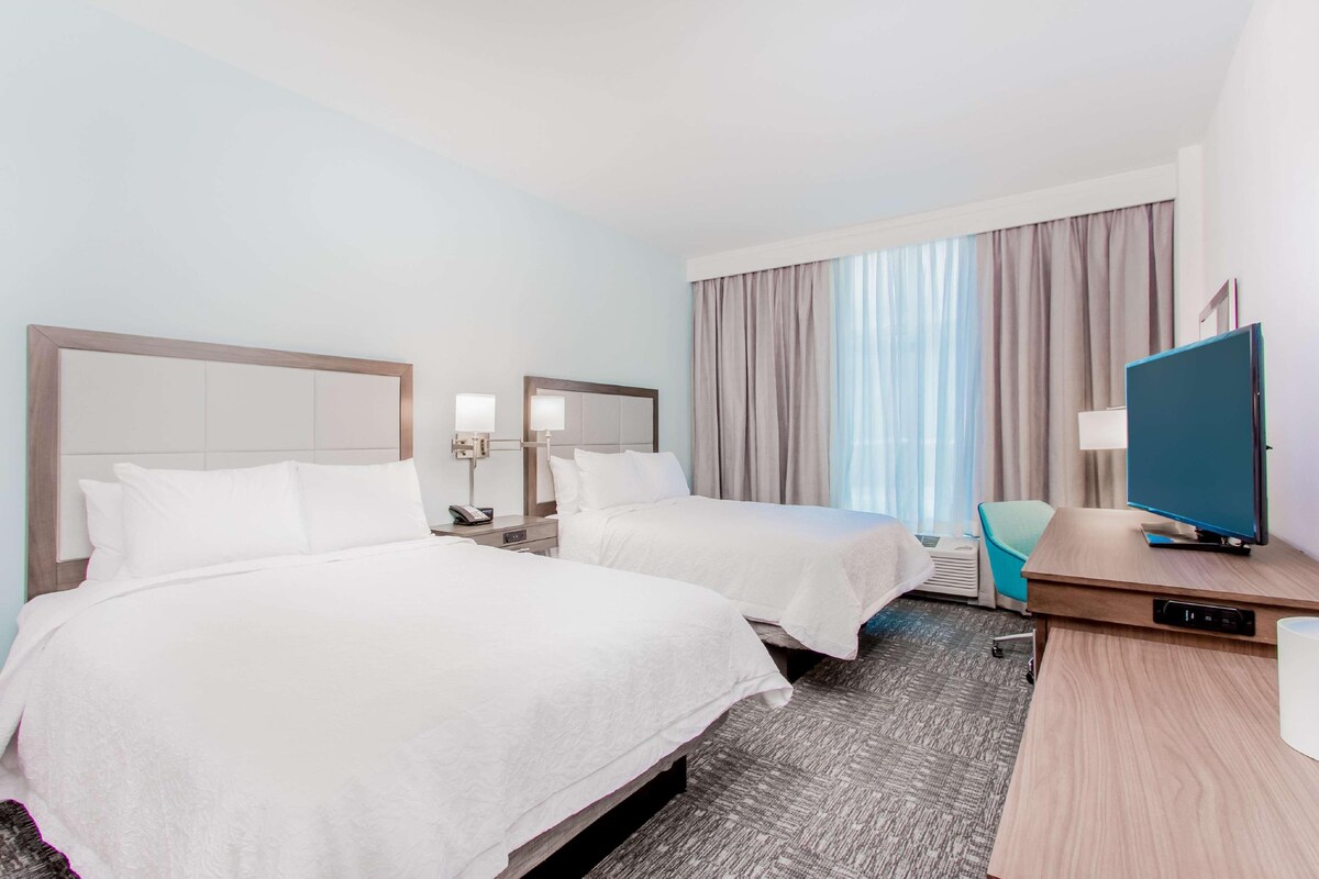 Una habitación de hotel con dos camas, un escritorio con televisor, una silla y una ventana con cortinas.