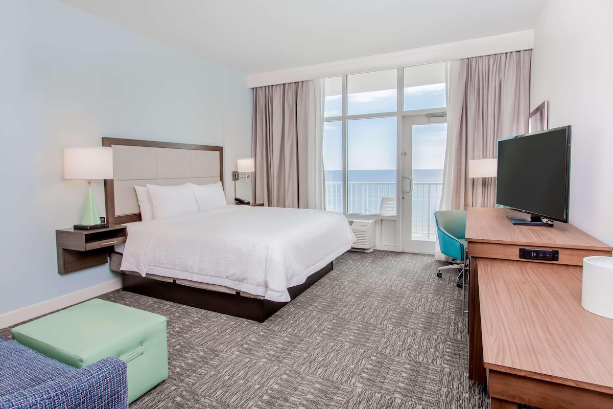Habitación de hotel con una cama grande, un escritorio con televisor, una silla, una lámpara y vistas al mar.