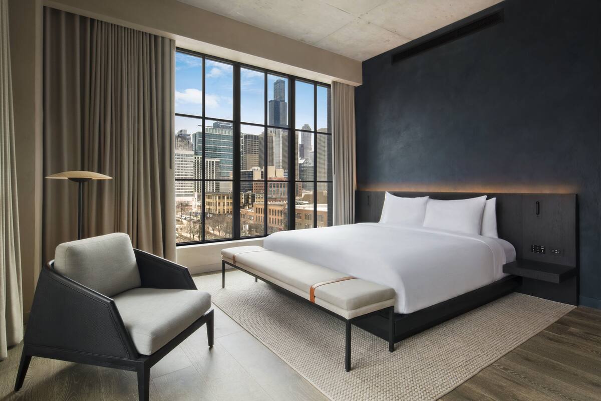 A modern hotel room with a large bed, a bench, a chair, and a view of the cityscape through large windows.
