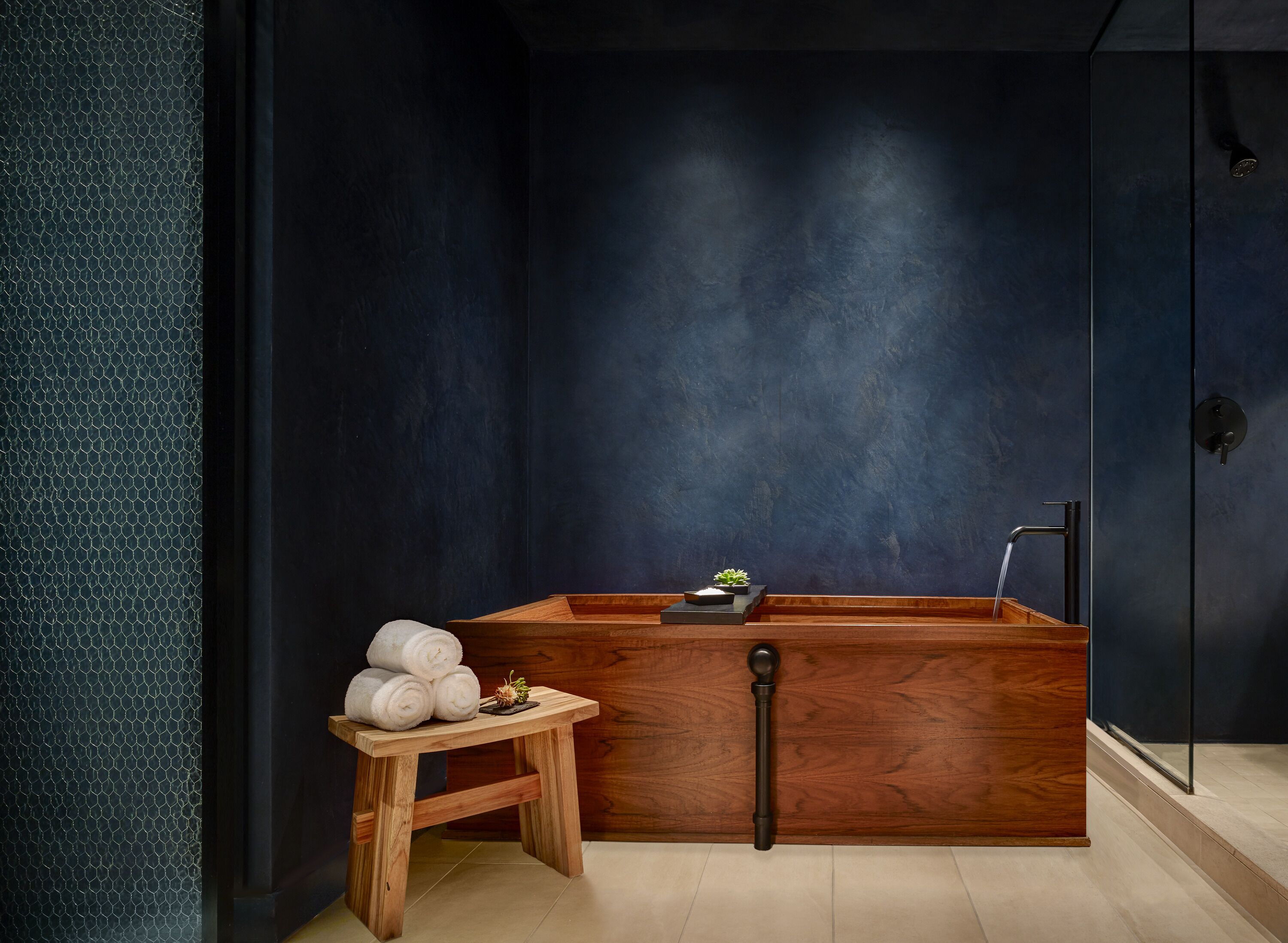 A modern bathroom with a dark blue wall, a wooden bathtub, a small wooden stool with towels, and a glass shower enclosure.