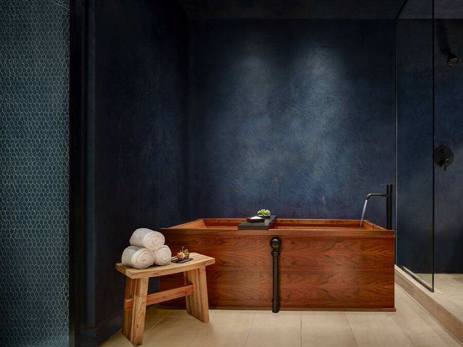 A modern bathroom with a dark blue wall, a wooden bathtub, a small wooden stool with towels, and a glass shower enclosure.