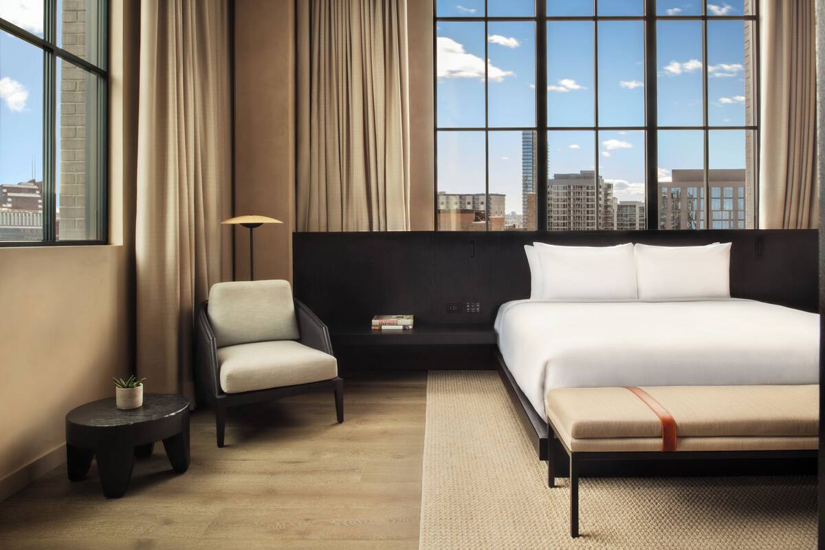 A modern hotel room with a large bed, a chair, a small table, and a view of the city through large windows.