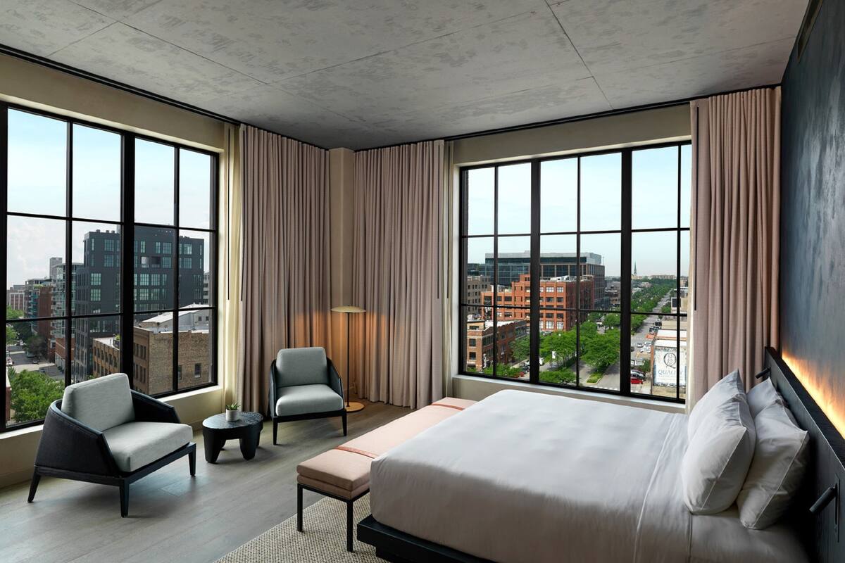 A modern hotel room with a large bed, two armchairs, a small table, and a view of the cityscape through large windows.