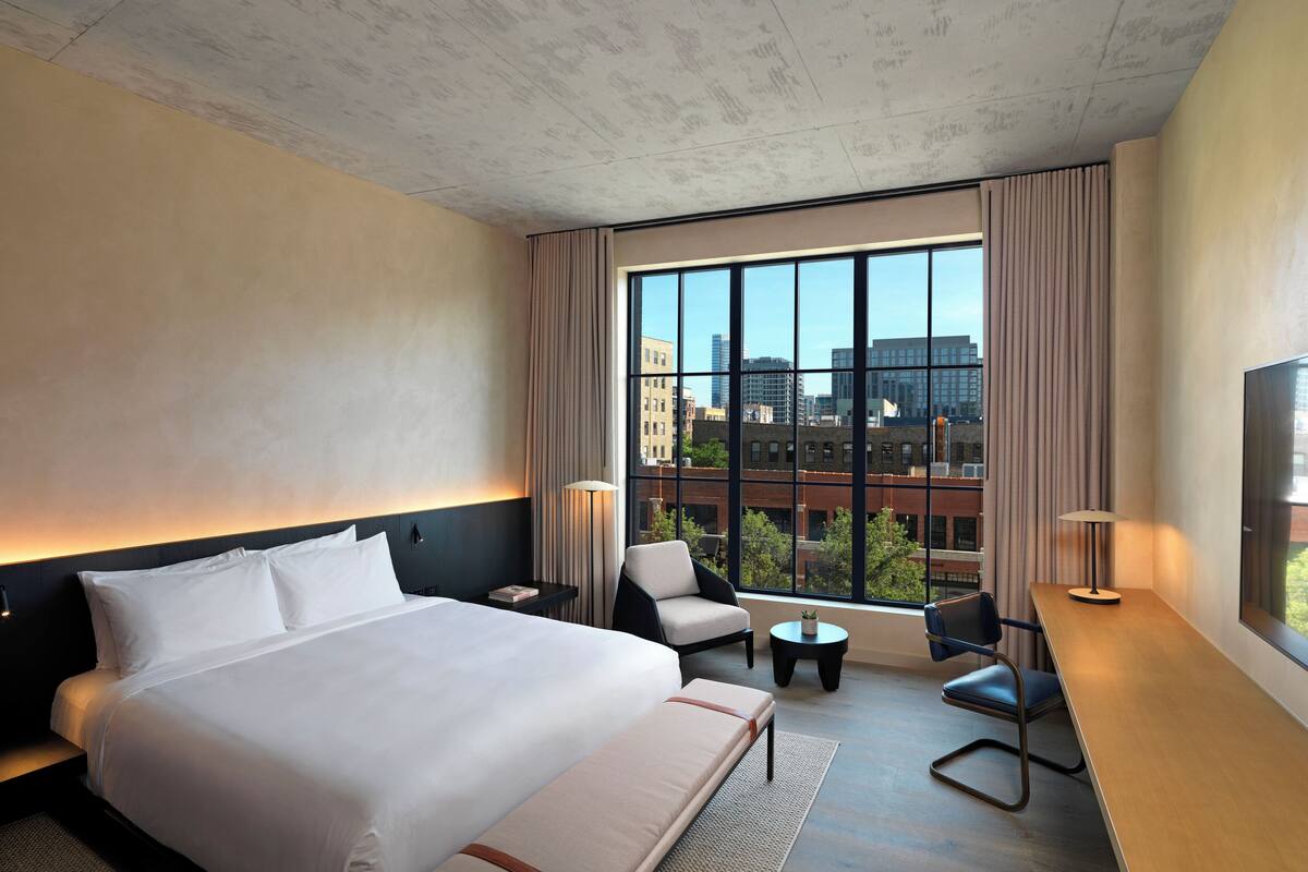A modern hotel room with a large bed, a desk, two chairs, a small table, and a large window offering a city view.