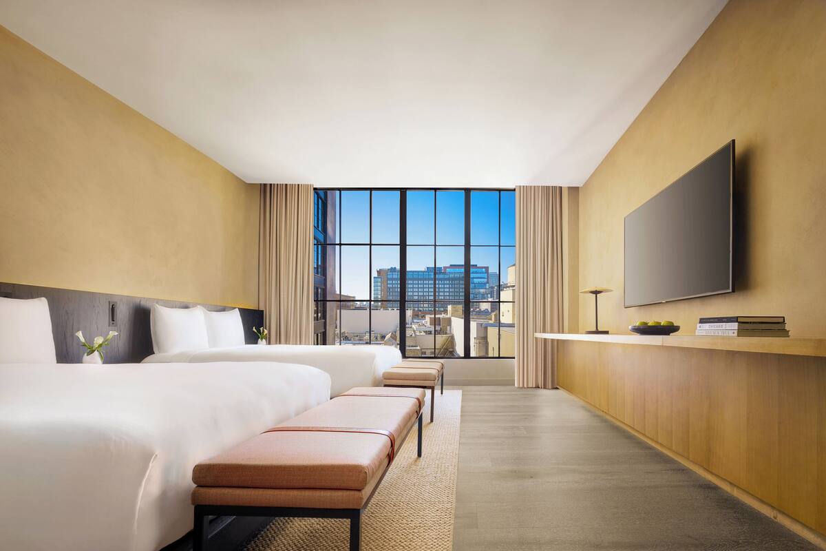 A hotel room with a large bed, a smaller bed, a bench, a TV, and a view of the city through large windows.