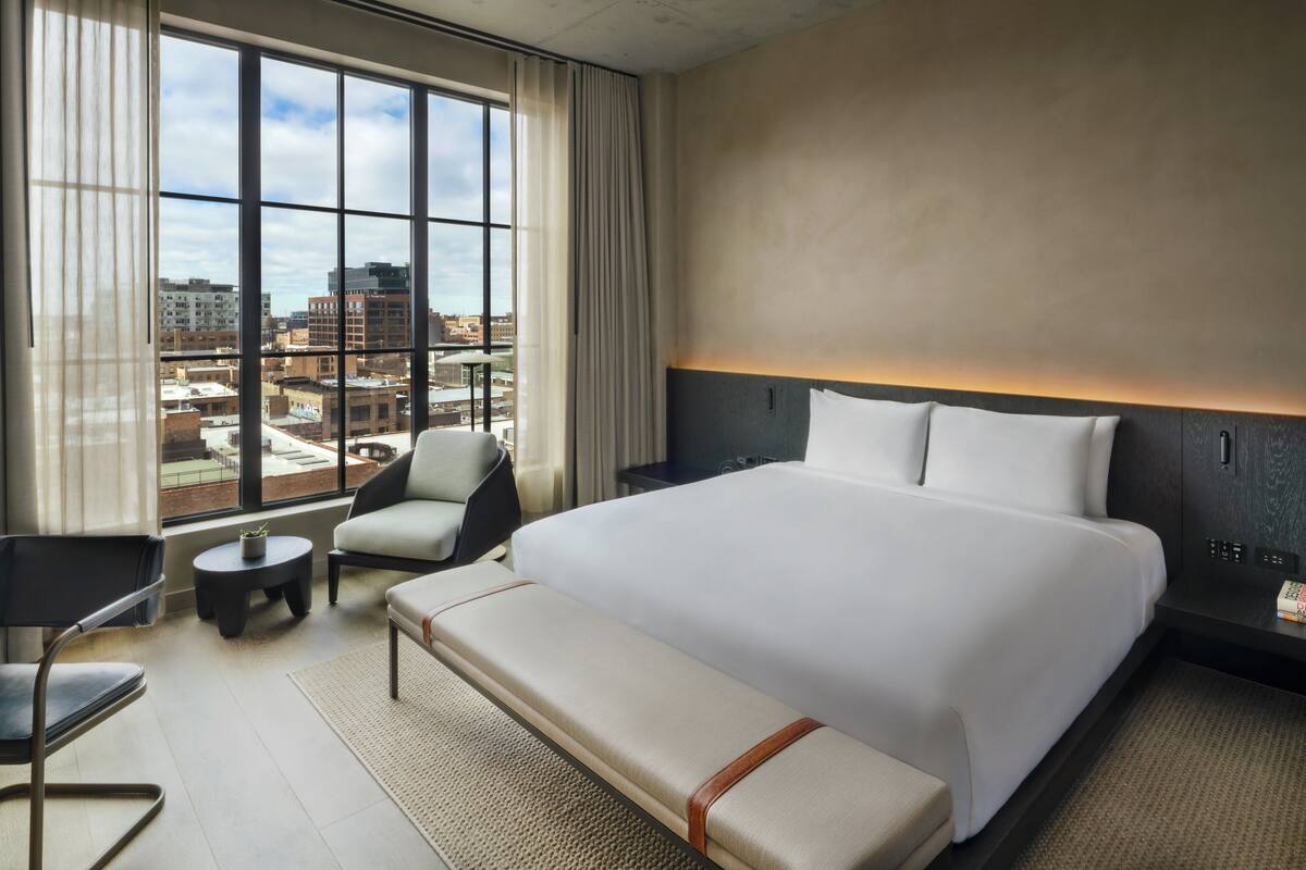 A hotel room with a large bed, a seating area with a chair and a small table, a nightstand, and a view of the city through large windows.