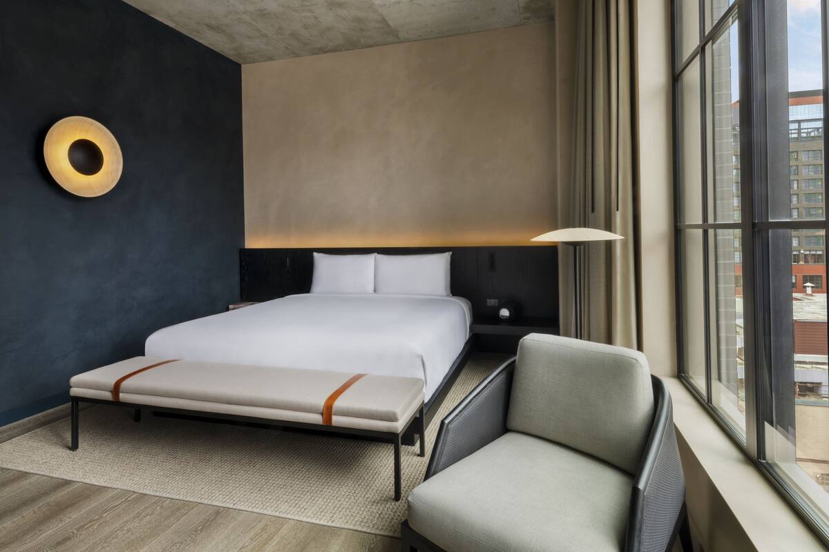 A modern hotel room with a large bed, a bench, a chair, a bedside table, and a large window offering a view of the city.