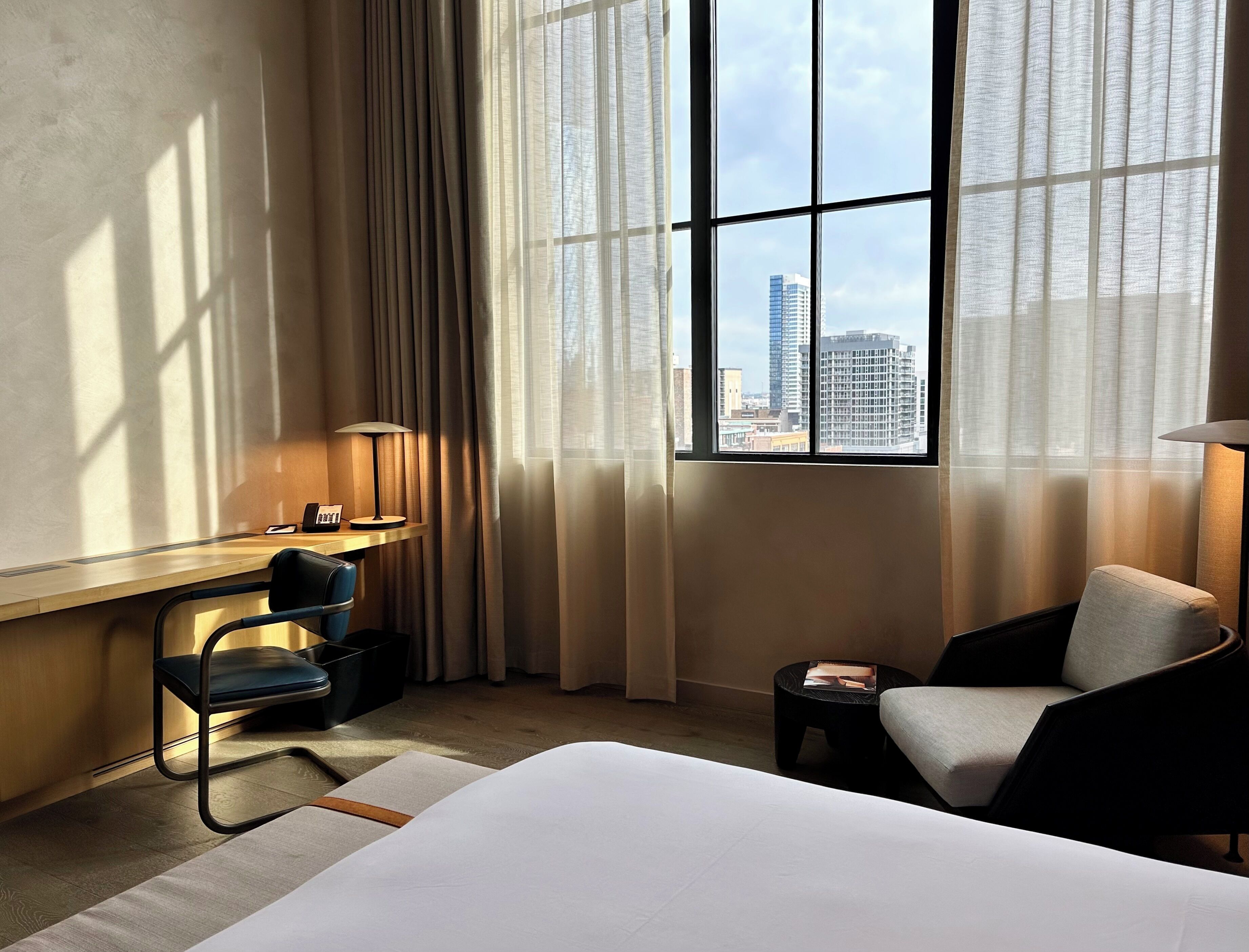 A hotel room with a bed, a desk with a chair, a lamp, and a view of a cityscape through large windows.