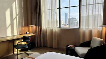 A hotel room with a bed, a desk with a chair, a lamp, and a view of a cityscape through large windows.