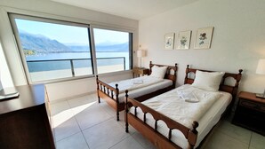 A hotel room with two wooden beds, a desk, and a large window offering a view of a lake and mountains.