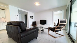 A modern living room with a black leather sofa, a wooden-framed rocking chair, a glass coffee table, and a flat-screen TV.