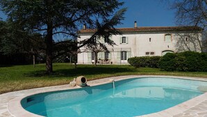 Una piscina con borde de piedra, un árbol grande y un edificio con techo de tejas al fondo.