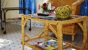 A wicker chair with a woven basket, a table with magazines, a remote control, and a vase with flowers. - Ose Cottages (Kisumu)