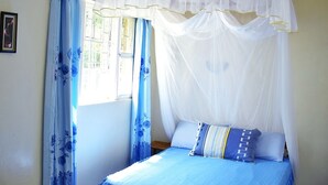 A bedroom with a bed under a white canopy, blue curtains with floral patterns, and a window with white frames. - Ose Cottages (Kisumu)
