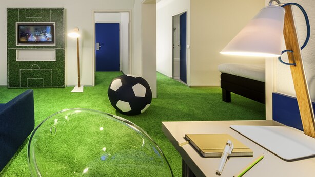 Een hotelkamer met een vloer van groen kunstgras, een grote zwart-wit voetbal, een bed, een bureau met een lamp en een aan de muur bevestigde televisie.